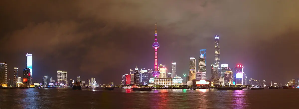 A colorfully lit nightscape panorama of the most faomous area in Shanghai. The area is surrounded by the Huangpu River, on which some cargo ships are driving. The peninsula is packed with skyscrappers. Most notably, the purple lit Pearl Tower, which has three large ball shapes. The second largest building in the world, the Shanghai tower with its twisting shape. And the Wold financial center, lit in blue with a square whole in its top.