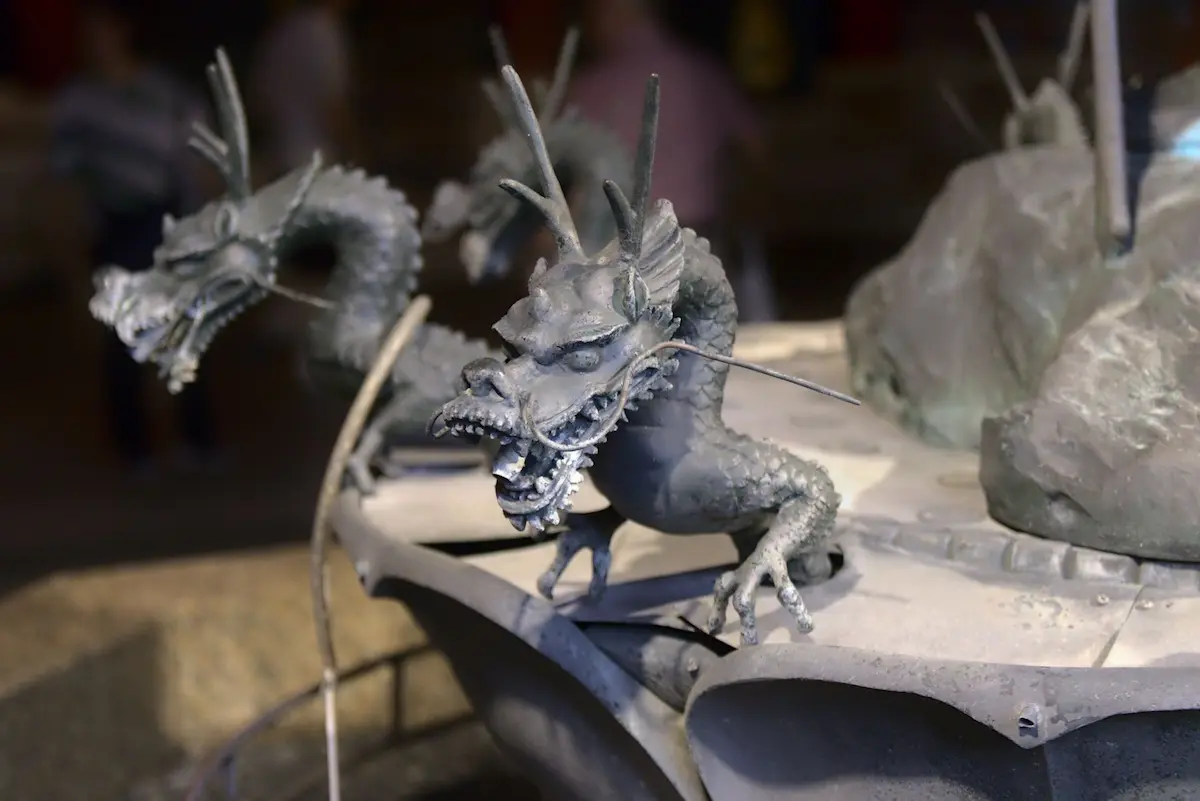 A dragon figurine above a water basin with a thin pipe in its mouth, usually spewing water, but now dry. There are several more of these in the background as they lign the round fountain.