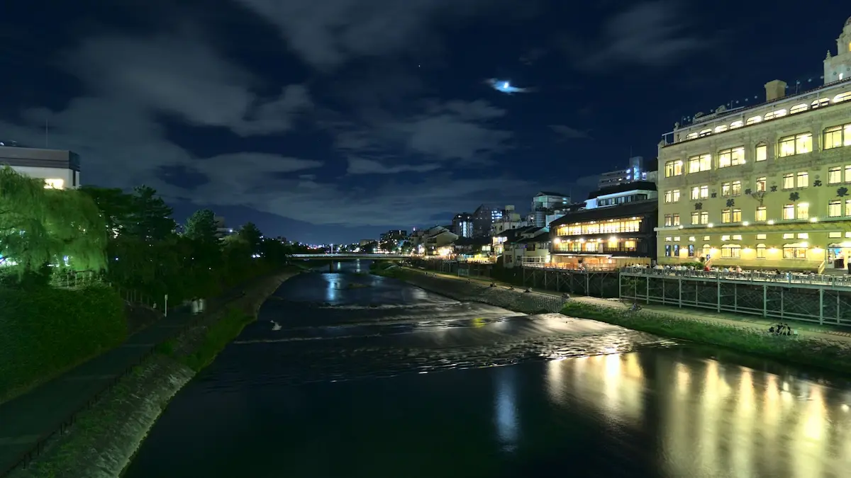 A long time exposure at night. The wide, but shallow Kamo river reflects the lights of restaurants, that are next to it. The waves and ripples in the water are smoothed due to the long exposure. A subtle crescent moon is hanging in the sky, illuminating a small cloud.