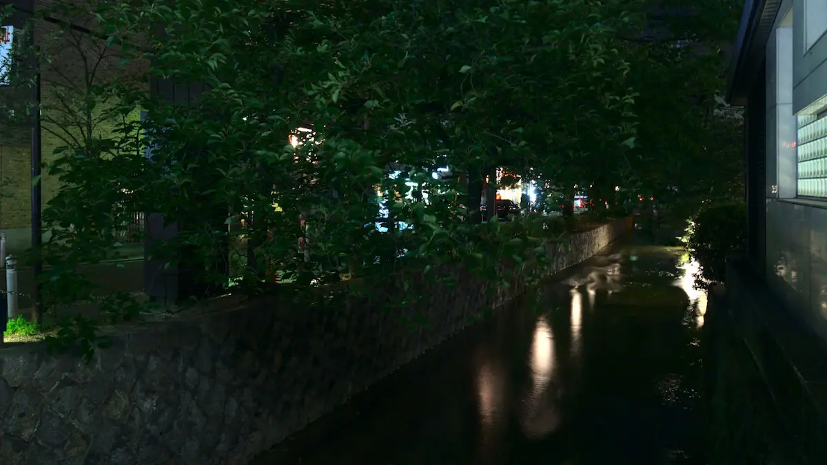 A nighttime long-time exposure. An about 3 meter wide canal, contained by a stone wall is the focus of the image. Lights reflect from its surface. Some trees are hanging over the canal from the side.