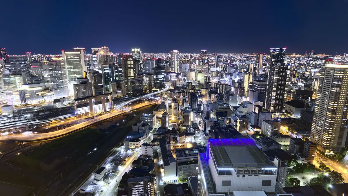 The nightscape of Osaka. In the bottom right, there is a helicopter Pad lit, with purple-blue lights. A bit street is making its way from the left to the middle, where a smaller street branches of, and vanishes into the urban jungle of Osakas never ending buildings.