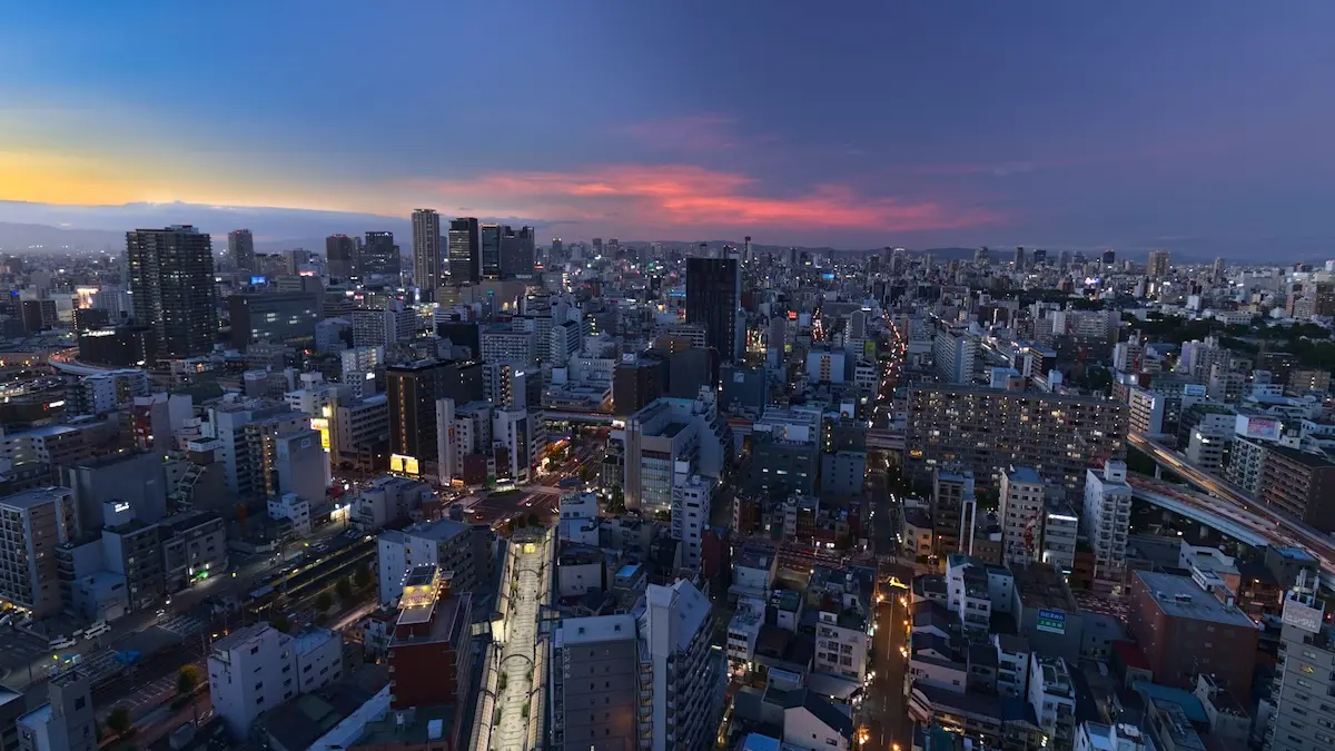 A view of Osaka during twilight. A few fluffy clouds are hanging in the sky and are illuminated bright red, by the sun below the horizon. The left part of the sky is glowing in a bright orange due to direct sun illumination. The lights of the city are slowly staring to turn on. Half an hour more and it will be night, turning the dense ocean of houses into a glowing wonderland, just waiting to be explored.