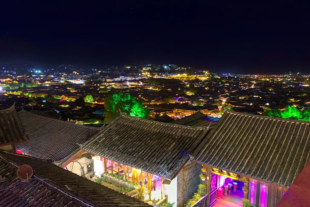 This image was taken from a rooftop bar. At the bottom there are two colorfully lit stores and/or bars in traditional buildings. Of the old town in the background some rooftops are lit.