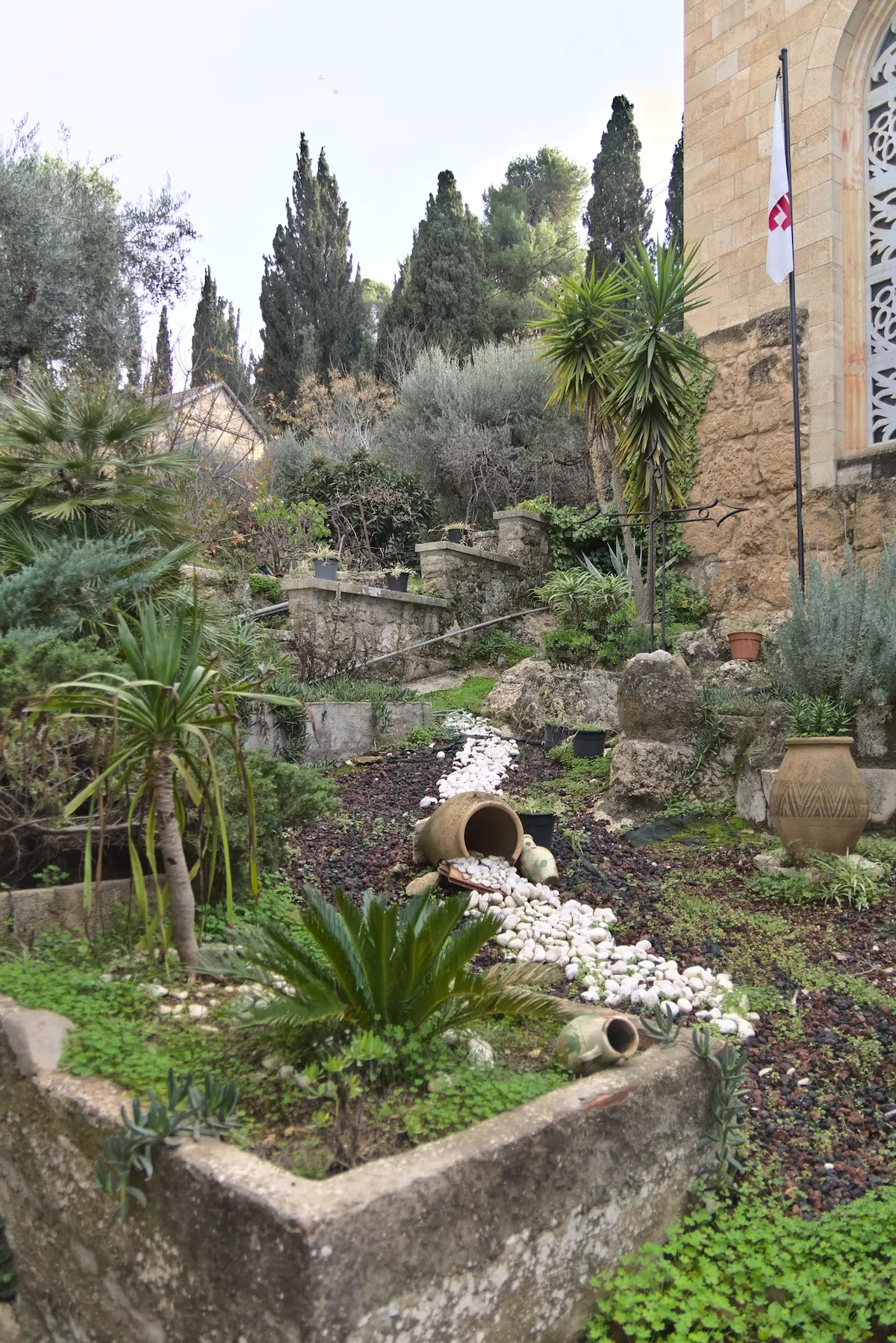 A stone garden surrounded by southern palm-like looking plants. The ground is made up of a mix of red and grey rocks. Some clay pots are intentionally placed, so that is looks like white rocks are spilling out of them.