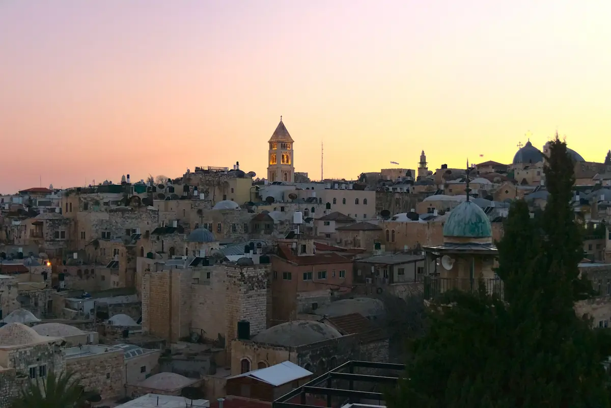 A view of the roofs of the old town of Jerusalem. The sky has an interesting purple-red-orange hue to it. In the center there is a church tower glowing from its illumination.