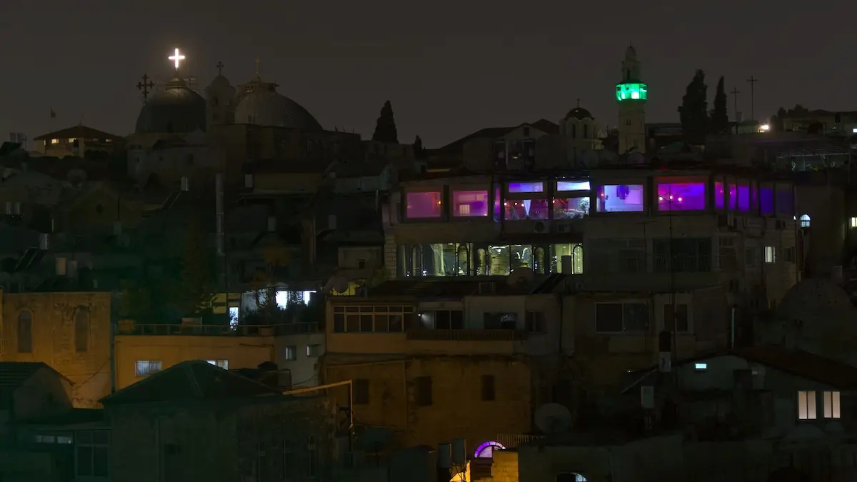 A night view of Jerusalem. In the right seems to be a night club, because of its colorful purple lighting. In contrast in the left is a church with a white lit christian cross.