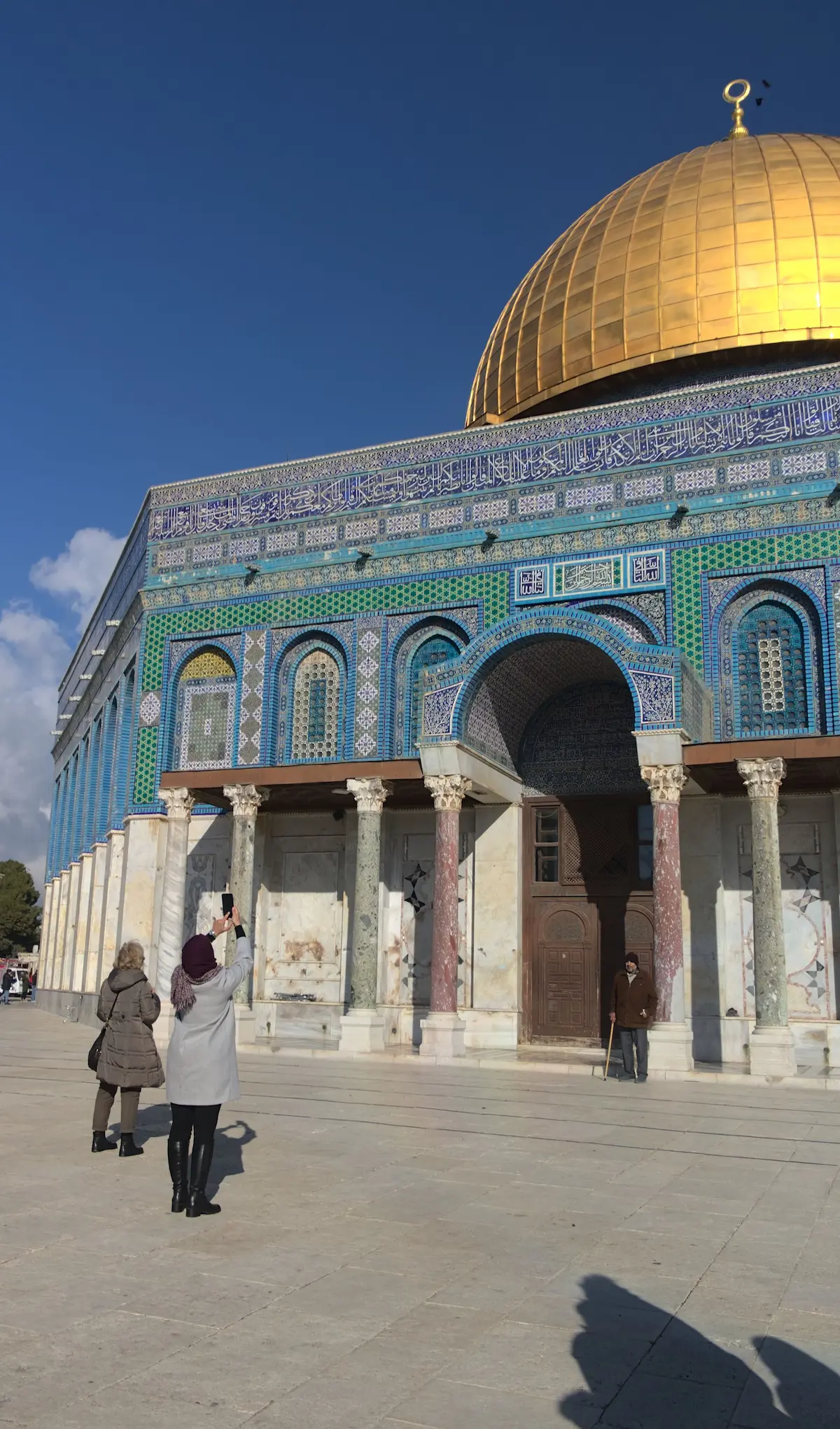 The Dome of the rock up close. Below the golden dome are the details of the wall well visible. Wich are intricate patterns in blue and green tones. In the foreground a woman is stretching to make a photo with her smartphone with an old man posing for the photo with a walking cane.