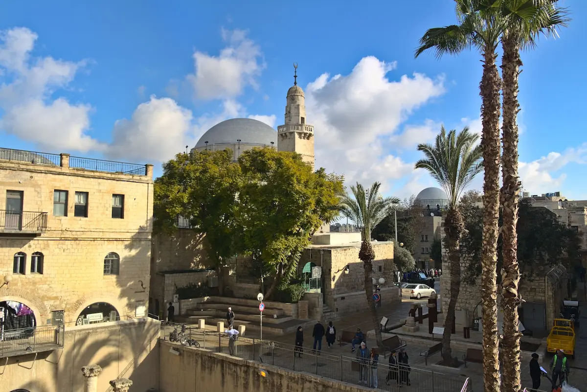 Some palm trees are stretching for blue the skies. a mighty tree in the center of the square down the stairs is giving shadow. The dome of a synagoge is visible above the tree in the backgrund. The streets look fairly busy with people, but not too crowded.